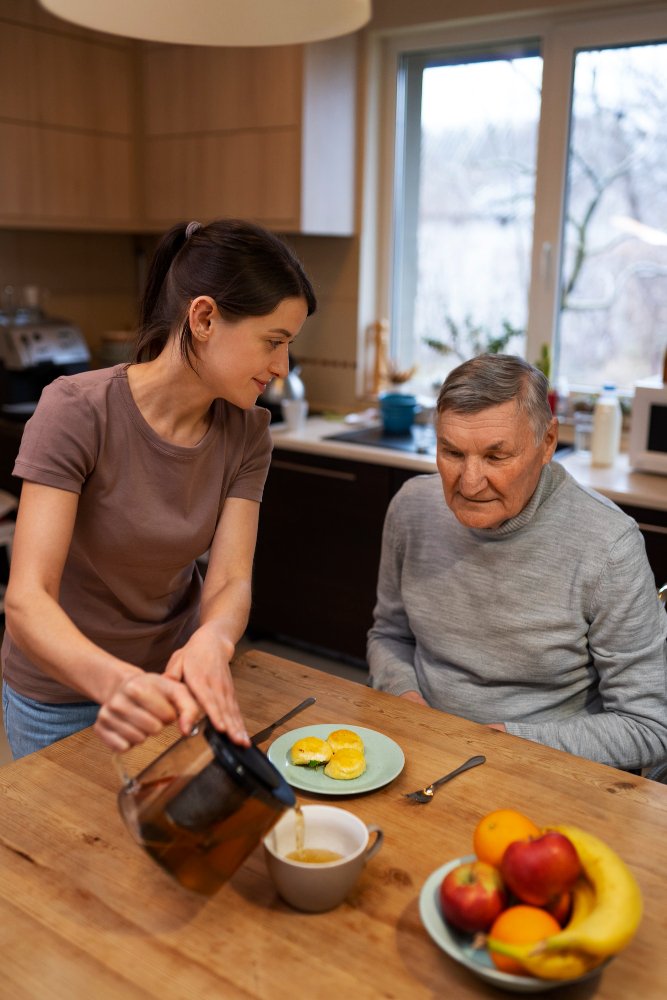 Adult Care and Support at Home A adult caregiver pours tea for an elderly man, providing home care assistance in a comfortable kitchen setting.