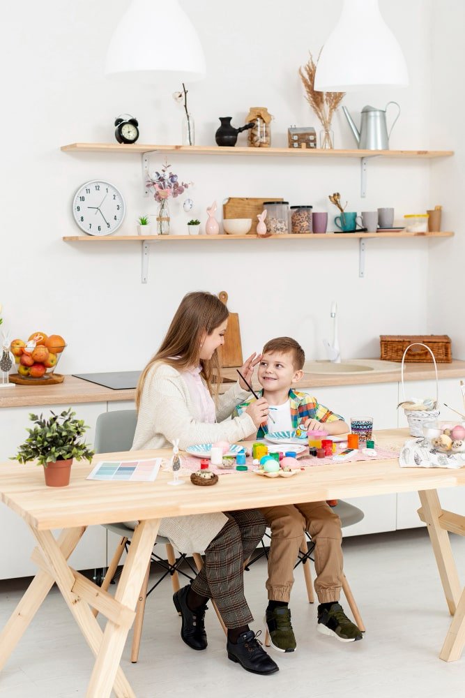 A carrer and boy are enjoying a fun, creative moment, painting Easter eggs together at a table with art supplies.