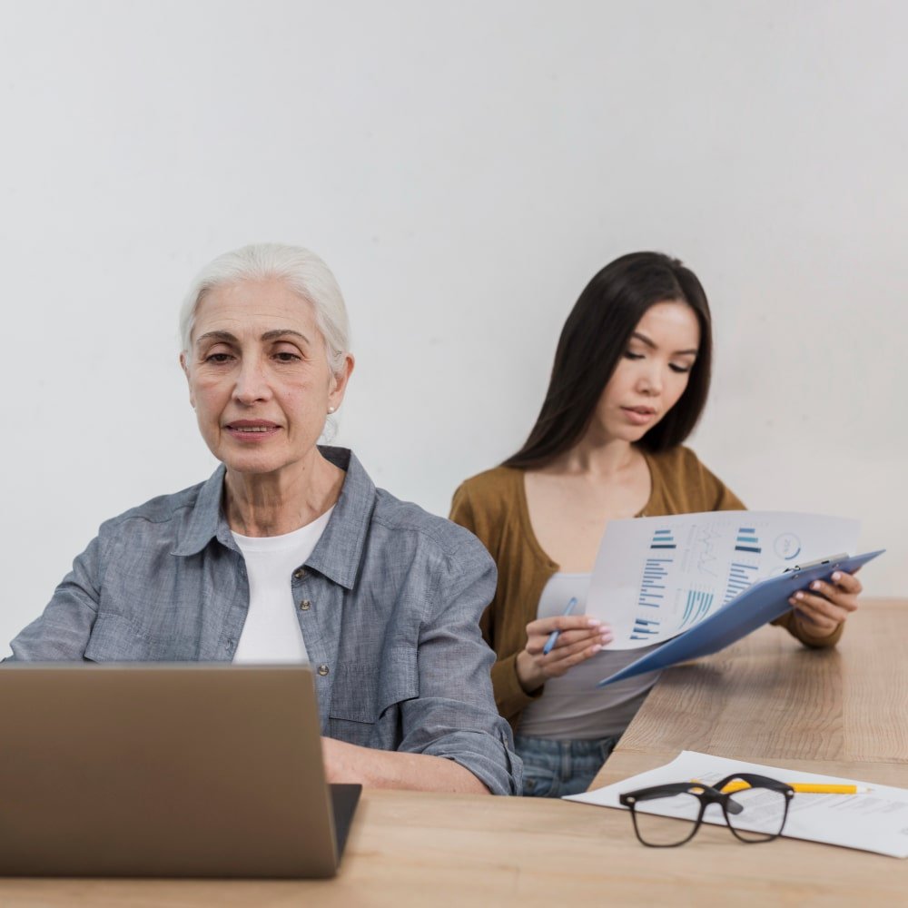 What You Can Expect From Joining Our Team An elderly woman and a young woman discuss documents in an office setting, reflecting the supportive work environment at Highbridge Care Solutions.